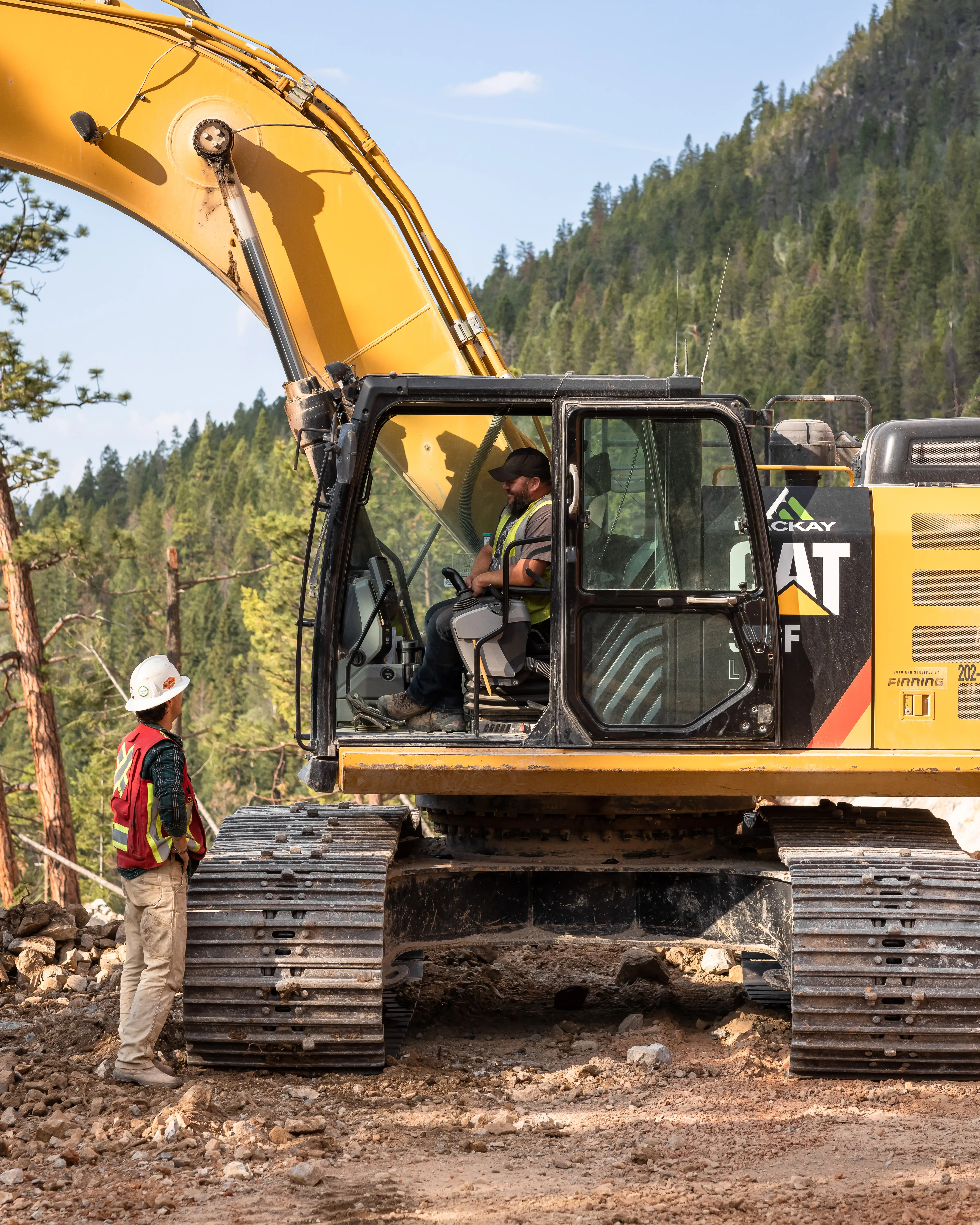 Mackay operator and spotter talking at an excavator on a forested mountain site